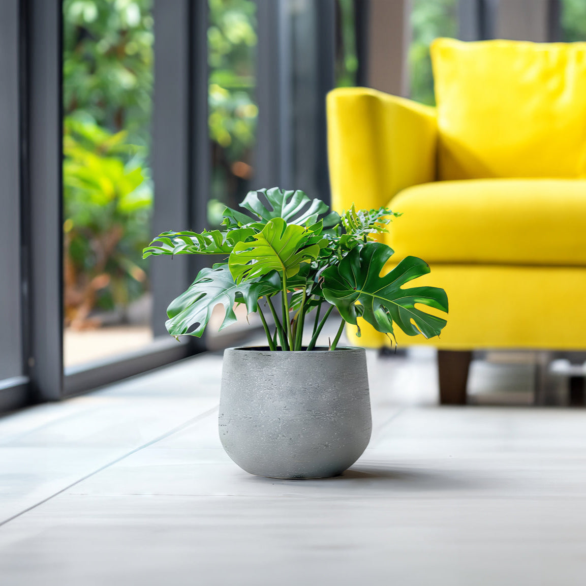 A lush monstera in a Fortezza Planter - Small from Landmark Decor sits on a light floor next to a bright yellow armchair. Large windows reveal the greenery outside, enhancing the luxury and natural beauty of the modern living space.