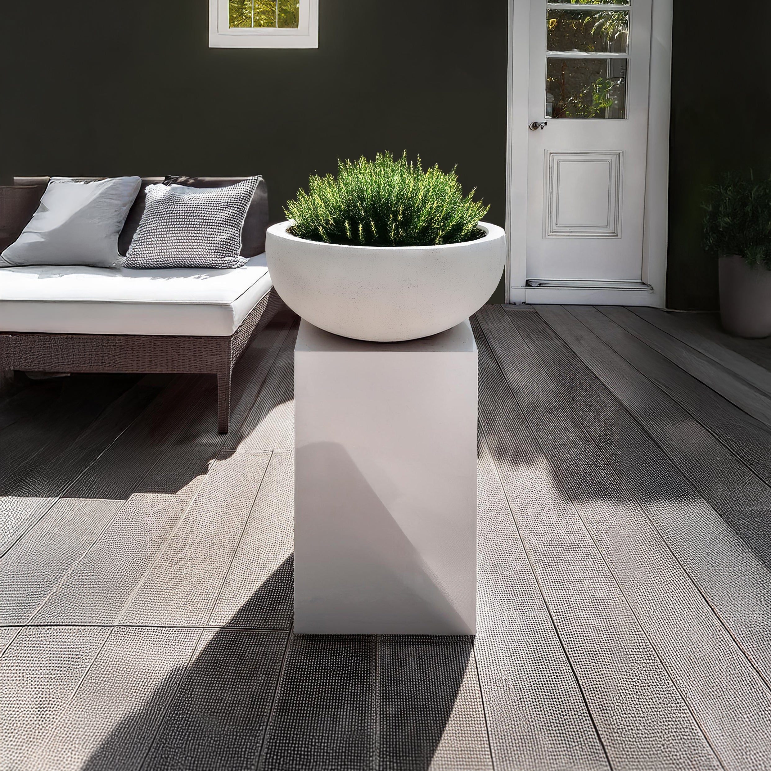 An Obelisk Planter by Landmark Decor with lush greenery sits on a tall white pedestal. The scene includes a white cushioned outdoor sofa, dark wall, and glass-paneled door, all complemented by textured wooden floorboards.