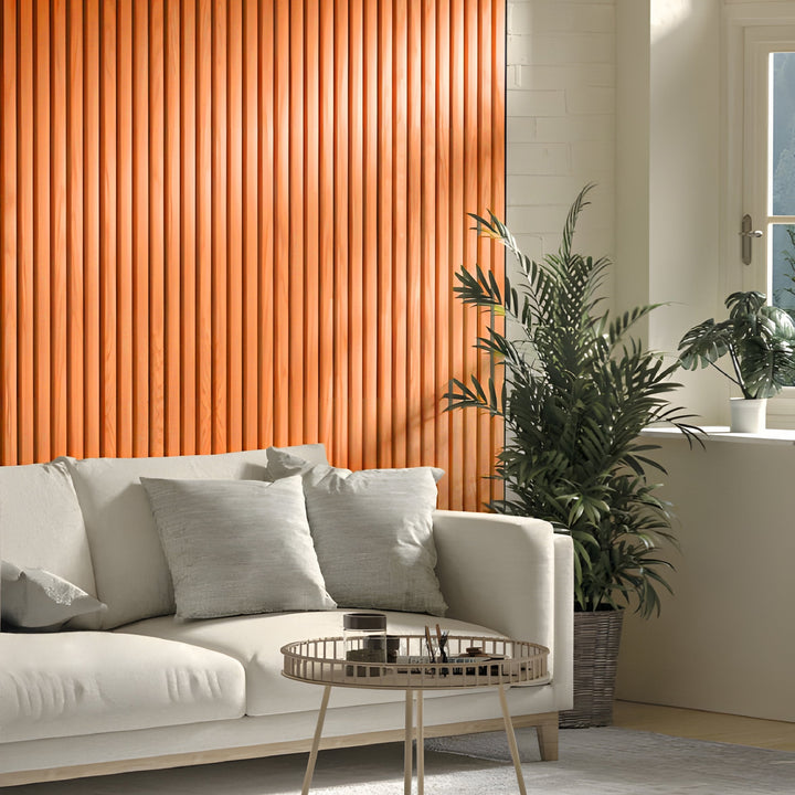 A modern living room with a beige sofa, light gray pillows, a round metal coffee table, potted plants, and an Orange WPC Fluted Slat Wall Panel by Landmark Decor accenting the sunlit window.