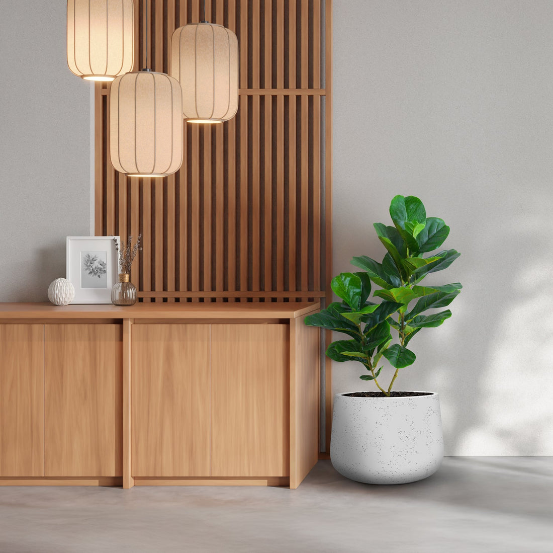 A minimalist room is styled with wooden cabinets adorned by Verdant Symphony from Landmark Decor, alongside a large potted fiddle fig and three lantern-style lights. A framed artwork and decorative vase enhance the cabinet against a wooden panel backdrop.