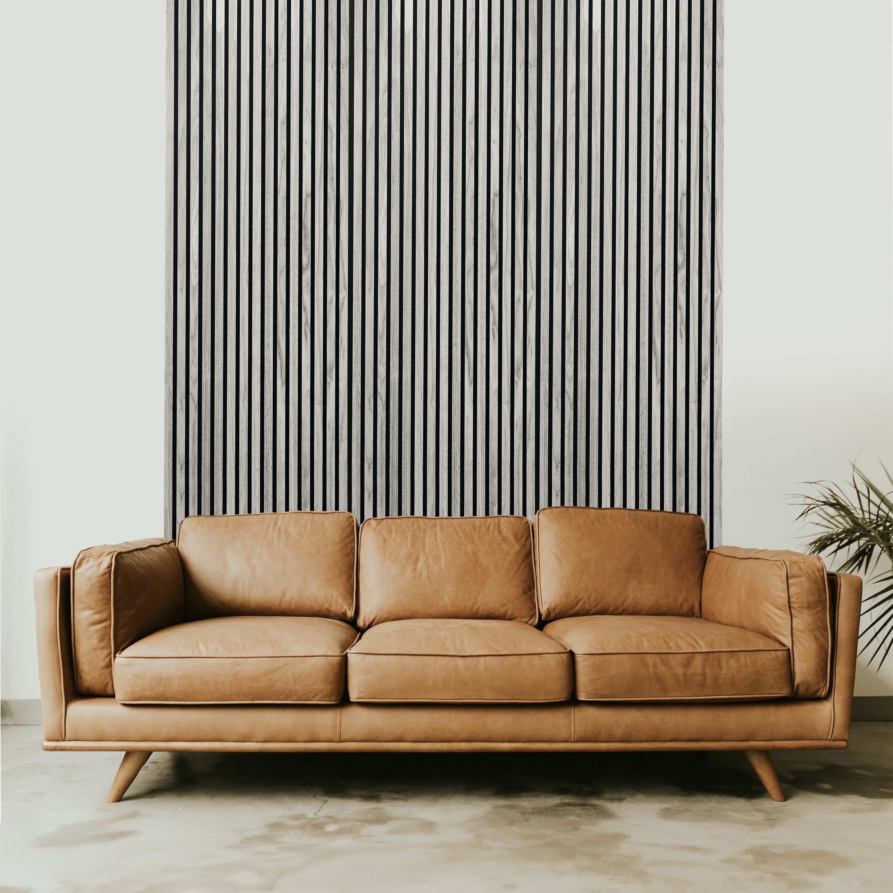 A tan leather sofa sits before Landmark Decor's Grey Oak Acoustic Slat Wall Panel, arranged vertically against a white wall, with a plant partly visible on the right for a modern design touch.