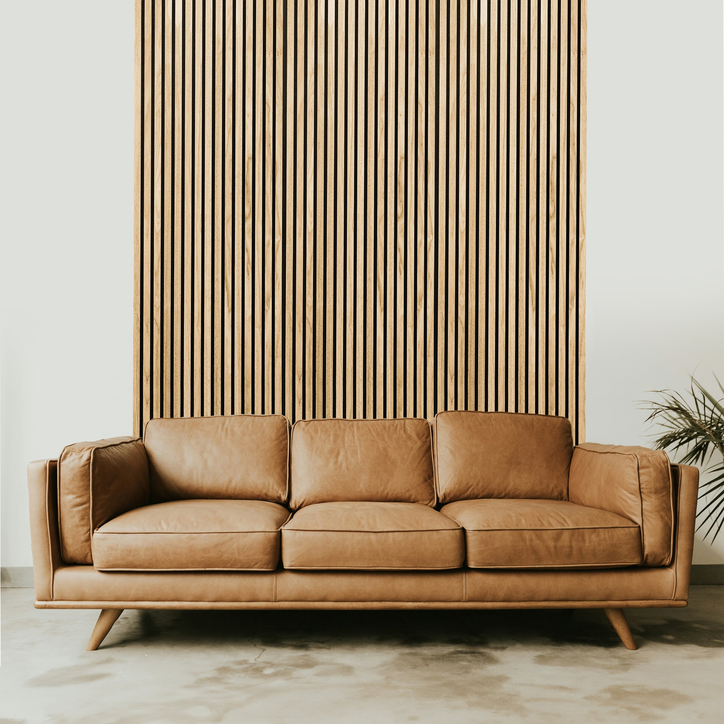 A brown leather sofa sits before Landmark Decor's Natural Oak Wood Color Acoustic Slat Wall Panel, which adds warmth and texture to the accent wall. A plant is partially visible on the right side.
