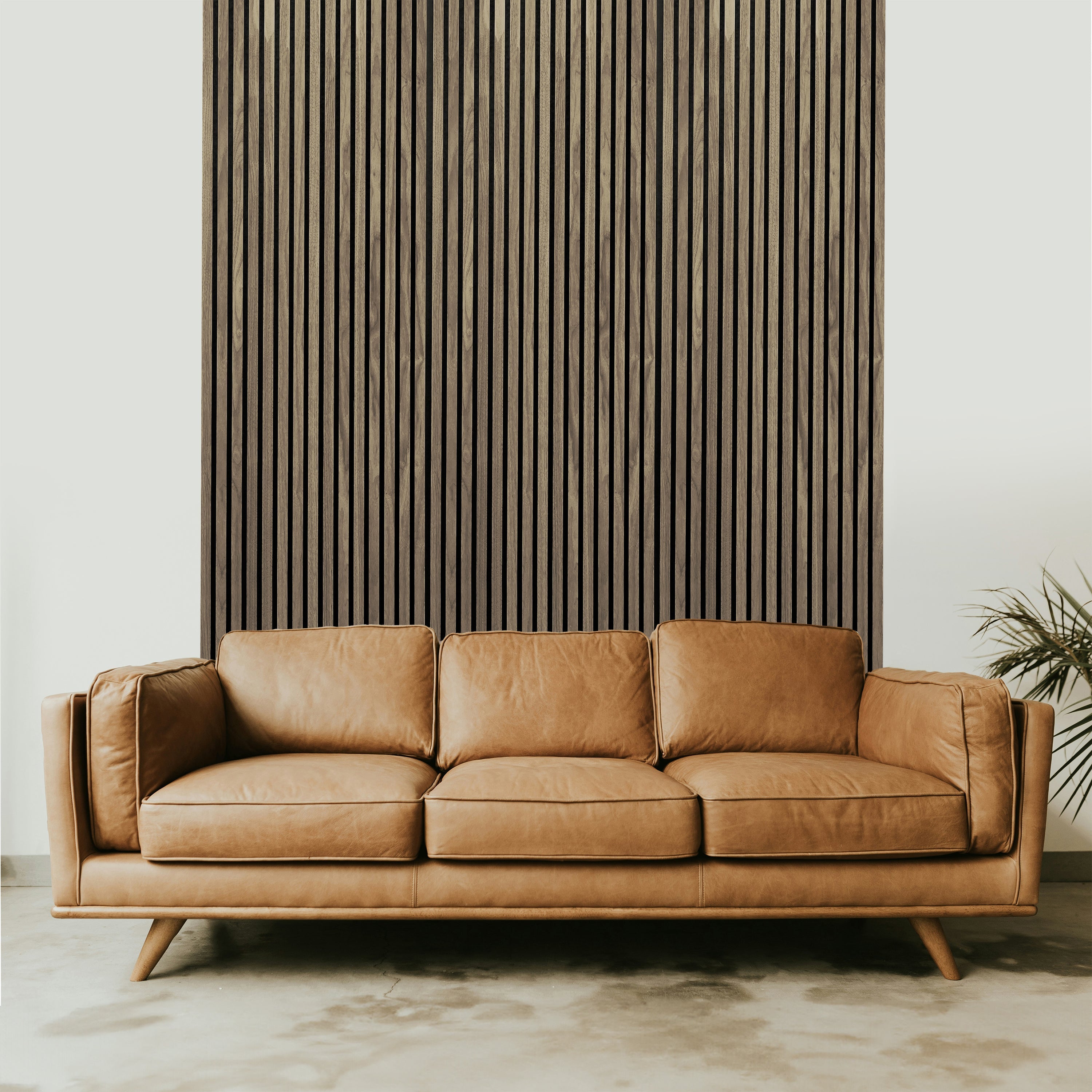 A tan leather sofa sits before a Landmark Decor Walnut Acoustic Slat Wall Panel, with a plant partially visible on the right, creating a stylish and elevated room design.
