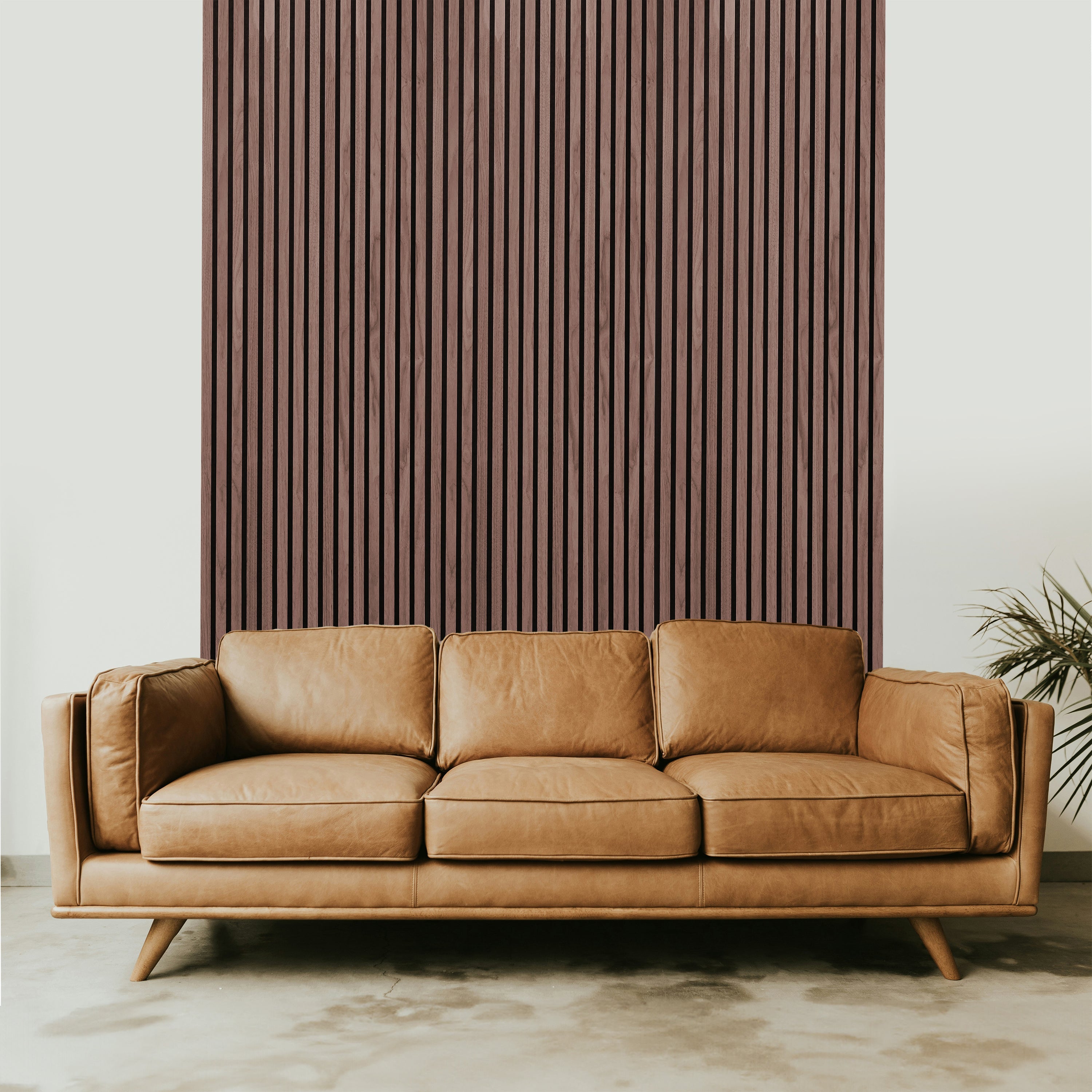 A tan leather sofa sits in front of Landmark Decor’s Dark Smoke Oak Acoustic Slat Wall Panel, arranged vertically as an accent wall, with a green plant partially visible on the right, adding warmth and style to the space.
