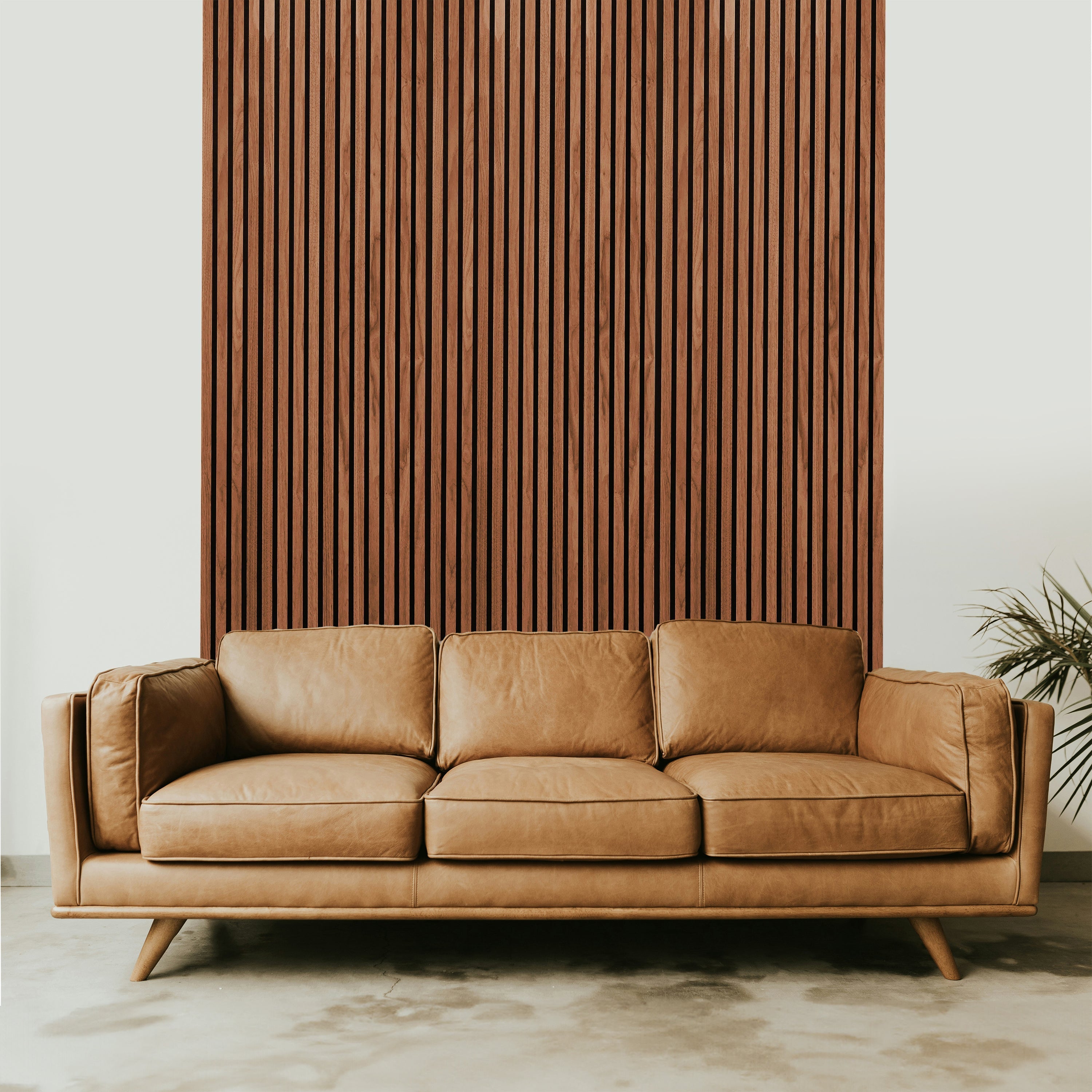 A tan leather sofa sits before a Landmark Decor Light Walnut Acoustic Slat Wall Panel, which brings real wood texture and soundproofing to the space. A lush plant is partially visible on the right side.