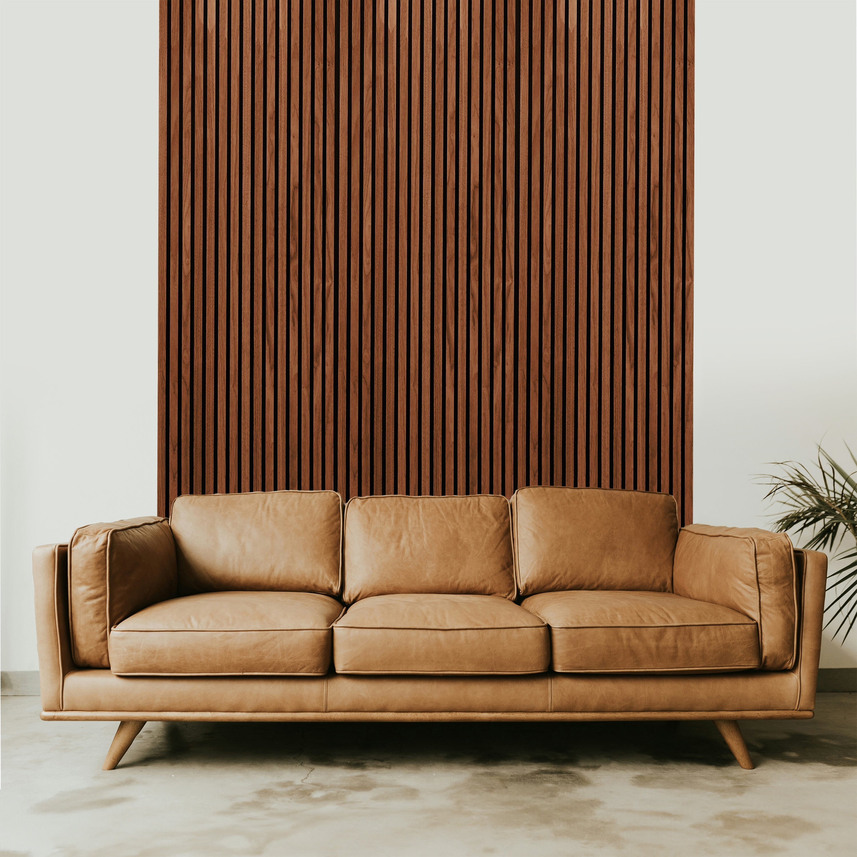 A tan leather sofa sits on a concrete floor in front of Landmark Decor’s Dark Walnut Acoustic Slat Wall Panel, with a plant partially visible on the right.